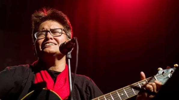 Shayan singing into a microphone with an acoustic guitar under red stage lights